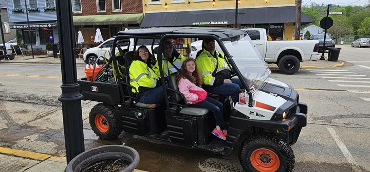 Volunteers in high-visibility jackets ride a utility vehicle down a small-town main street